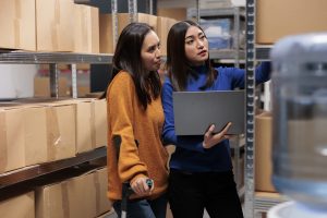 Warehouse managers preparing customer order and checking pick ticket on laptop. Storehouse young asian women employees searching parcel on shelf while using inventory software